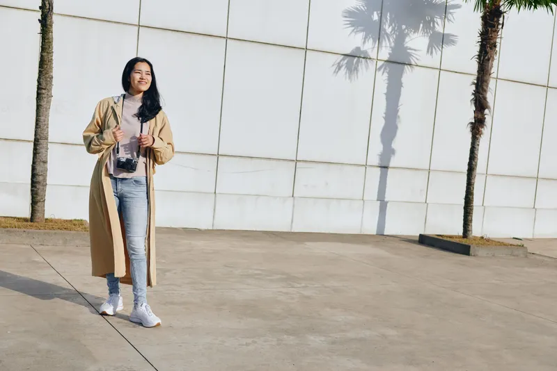 Woman in beige coat standing outdoors beside a white wall, daylight enhances casual fashion with denim and white sneakers, street style and confident vibe