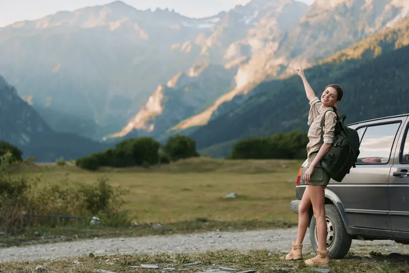 A young woman with a backpack stands beside a car in a rugged mountain landscape, waving happily.