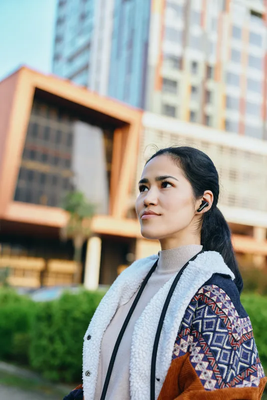 Woman outdoors in city architecture, wearing jacket and scarf, thoughtful expression, modern buildings in the background, daylight, urban fashion lifestyle vibe