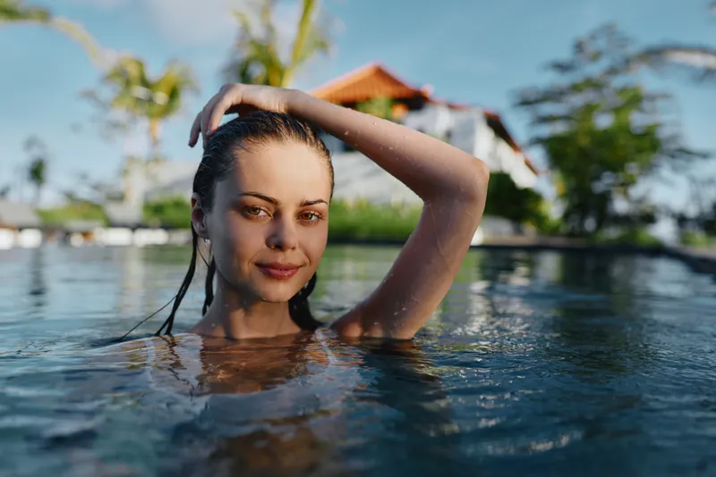 woman pool water leisure in tropical resort, smiling serene, standing in clear pool with hands on head, bright sunshine, palm trees and villas in background, relaxed lifestyle moment