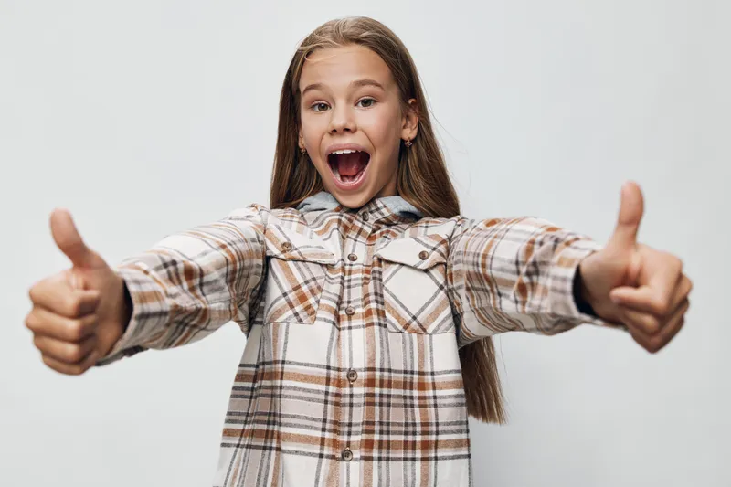 Girl with long hair wearing plaid shirt shows thumbs up with excited face expression standing isolated on white background. Positive emotion and happiness concept.