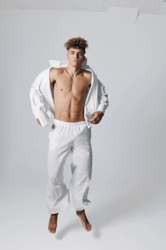 Young man with curly hair wearing white pants and open white shirt standing barefoot on white background in studio with confident expression and relaxed posture.