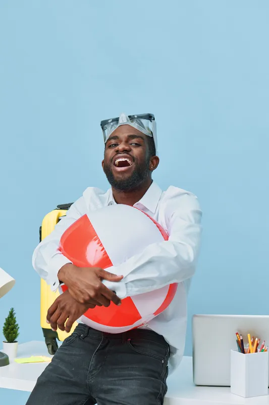 Fun and energetic young man with dark skin and cheerful expression holding a beach ball in a modern office environment with blue background. Casual style, positive mood, lifestyle concept, vibrant col