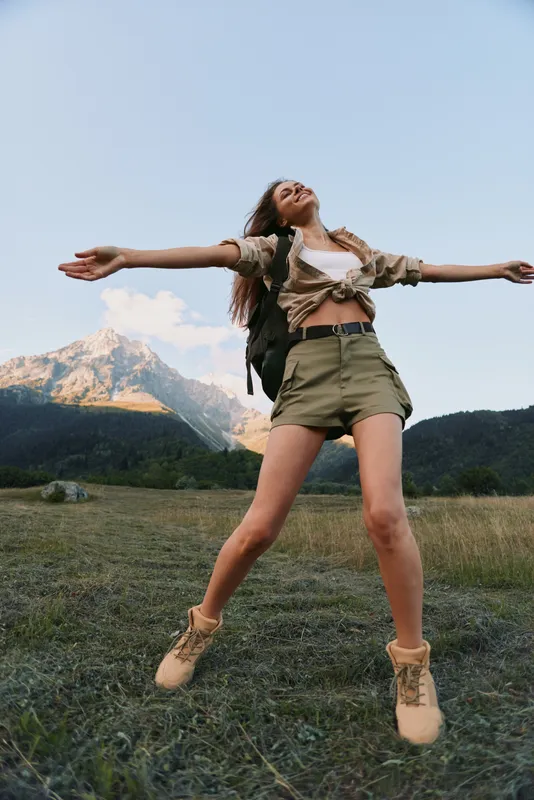 A woman leaps in a grassy field with distant mountains, capturing a carefree outdoor lifestyle, happiness, and a sense of freedom in a sunlit landscape. Bright sky, vibrant energy.