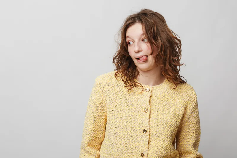 Young woman with curly hair wearing yellow knitted sweater looks sideways with playful expression and tongue out. Studio portrait on light grey background. Concept of fun and spontaneity.