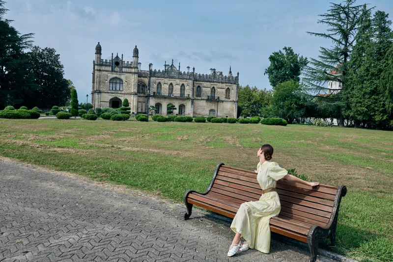 Woman sits on a wooden bench in a sunny park, with a historic building in the background, conveying calm leisure and heritage discovery.