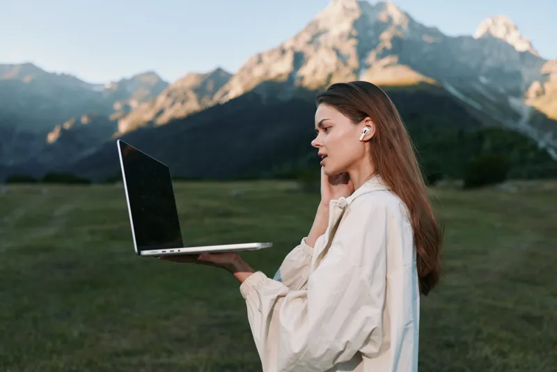 A focused woman stands in a scenic meadow, holding a laptop with one hand while speaking on the phone, exuding concentration, confidence, and calm during outdoor work.