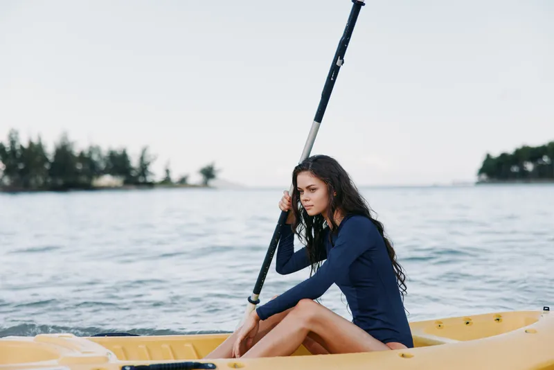 Young woman canoeing on a calm lake with trees in the background, wearing a navy long-sleeve shirt and focused expression.