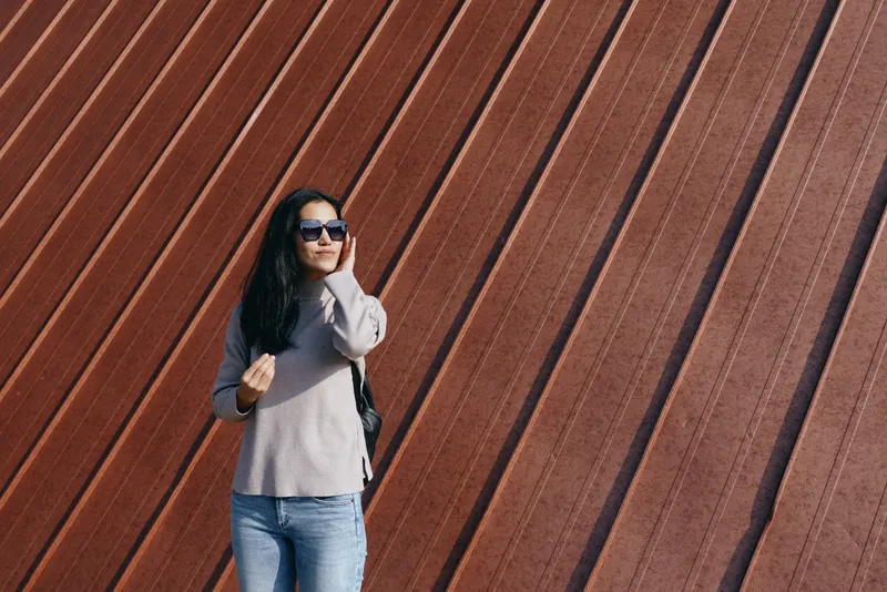 Street fashion with a woman wearing sunglasses, casual jeans and sweater, posed against a diagonal striped rust wall in an urban outdoor setting, contemporary vibe