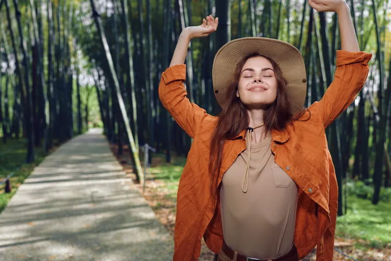 Woman hat forest path smile happiness travel nature portrait outdoors with arms raised, serene relaxed lifestyle moment, autumn sunlight on trees, peaceful female enjoying nature journey.