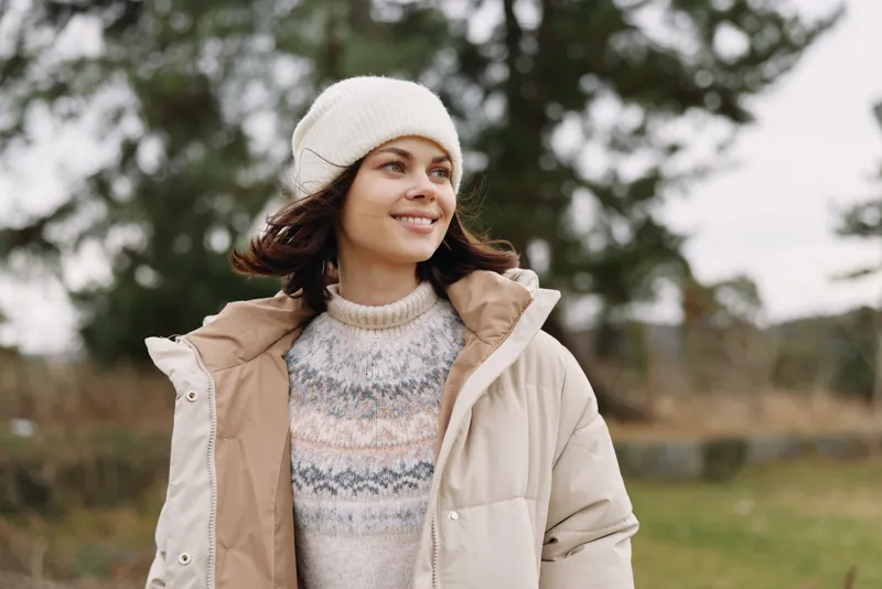 Woman in a warm coat and beanie outdoors, smiling in a grassy field with trees, capturing a cozy, natural lifestyle moment in cool weather.