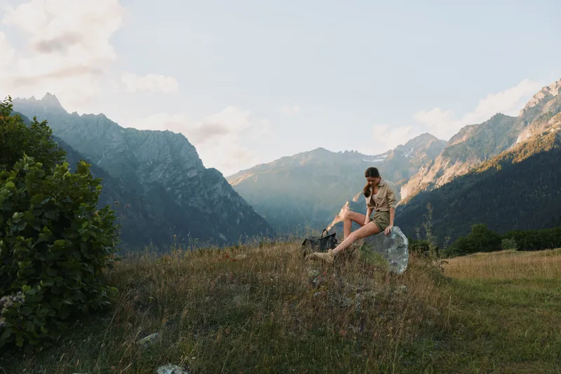 Woman enjoying outdoor lifestyle in a mountain meadow, sitting on a rock and gazing at distant peaks, casual clothes, peaceful mood, warm sunlight, natural landscape and solitude inspires calm.