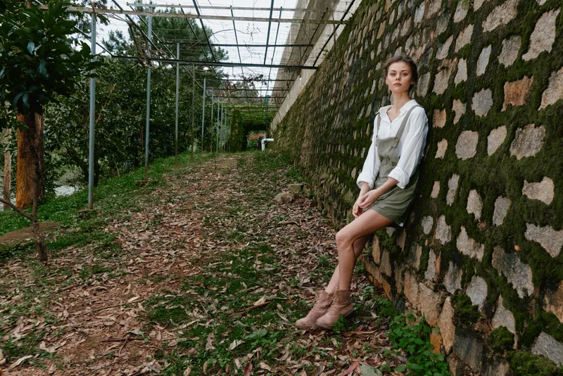Young woman in casual outfit sitting on stone wall surrounded by greenery, conveying a serene and thoughtful mood in a nature inspired setting
