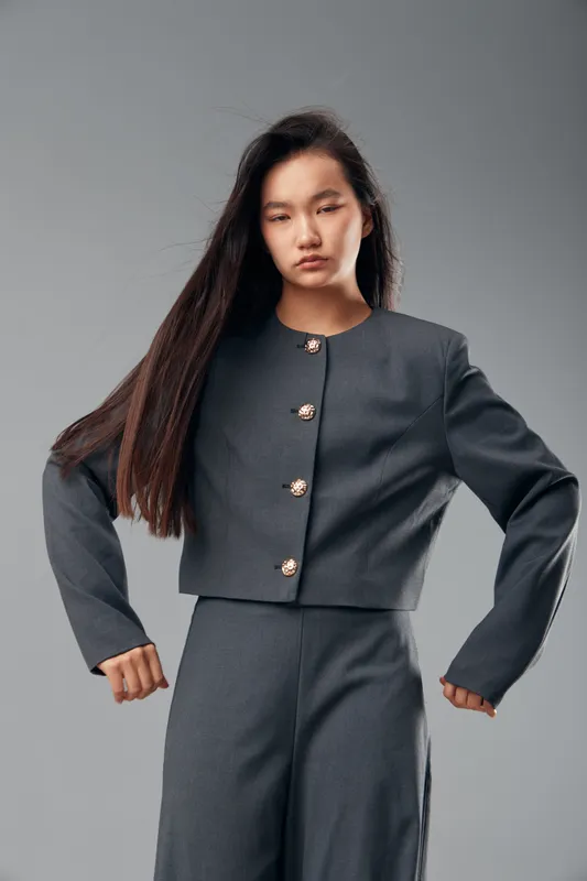 Young woman wearing gray tailored suit with long sleeves and decorative buttons posing with hands on hips against plain gray background for fashion portrait.
