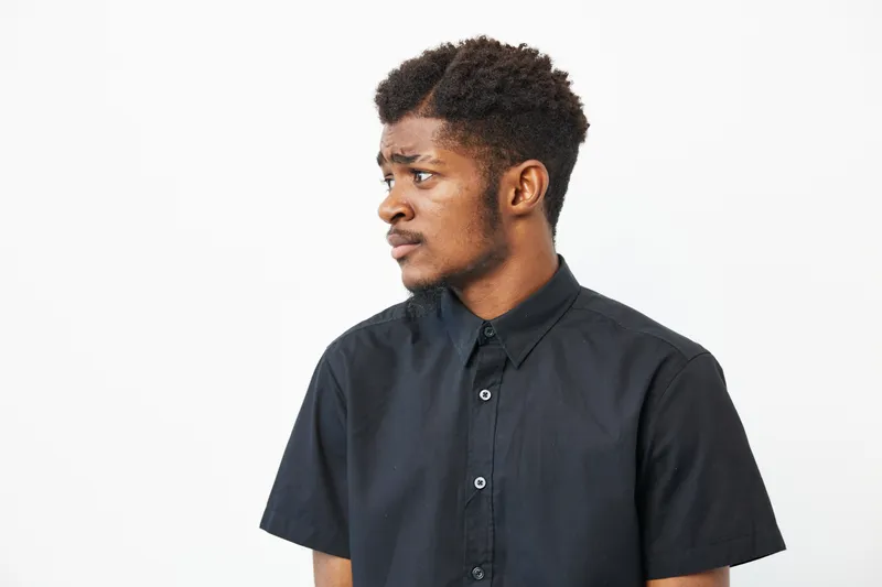 Black man posing thoughtfully in a stylish black shirt against a neutral background, conveying a sense of contemplation and confidence.