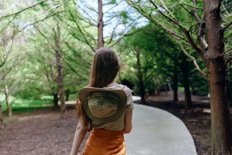 Woman walking on curved path through green trees, nature scene with hat hanging on her back, orange skirt and long hair, casual outdoor leisure and peaceful forest stroll