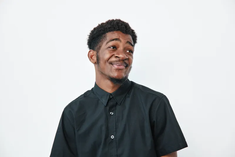 Young African American man with dark skin wearing black shirt looking sideways with smile on white background. Portrait, casual style and positive emotion for lifestyle concept.