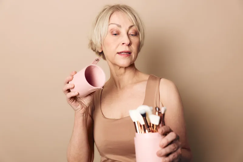 Mature woman with short blonde hair holding pink container and makeup brushes in beige top. Neutral background highlights beauty routine and self-care in natural light for skincare concept.
