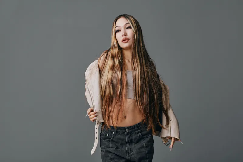 Young woman with long hair posing in stylish casual outfit with jacket over shoulder against gray background. Fashion, confident model with natural makeup in studio.
