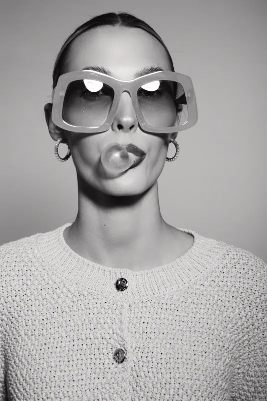 Young white woman with pale skin wears large sunglasses and knitted sweater while blowing a bubble with chewing gum.