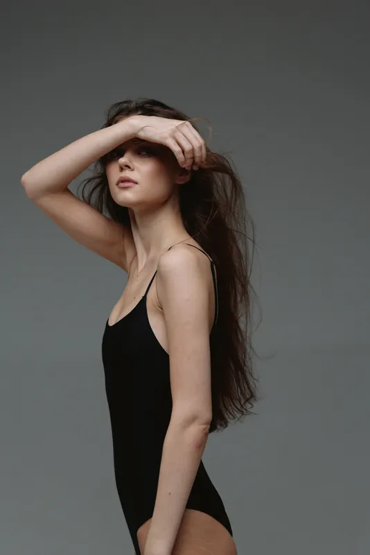 Young woman with long hair in black bodysuit posing with hand on forehead against grey background