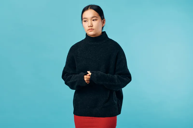 Woman standing with hands clasped wearing dark sweater and red skirt, posing against blue background. Female model with calm expression and casual winter outfit in studio.