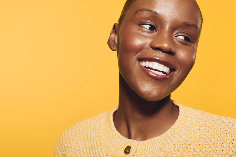Smiling African woman with dark skin wearing a yellow sweater looks away on a bright yellow background. Concept of happiness, beauty, and natural skin glow with warm lighting.