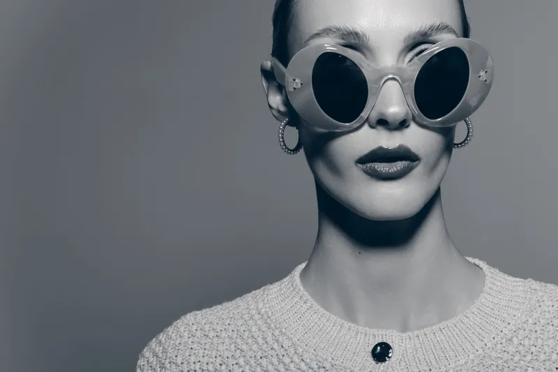 Portrait of a pale skinned Caucasian woman wearing round sunglasses and hoop earrings in a monochrome studio setting.