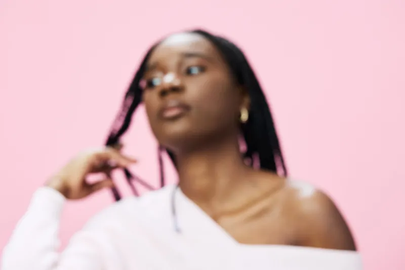 Young woman with braided hair, wearing a stylish off shoulder top, showcasing confidence against a soft pink background, emphasizing modern beauty and self expression