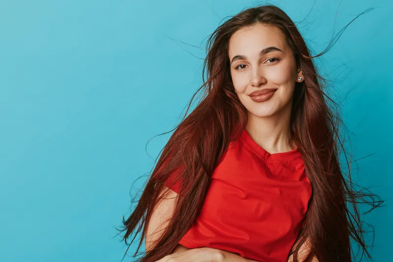 Young woman with long hair wearing red shirt smiling confidently against blue background. Portrait of happy female model with natural makeup and relaxed pose in studio setting.