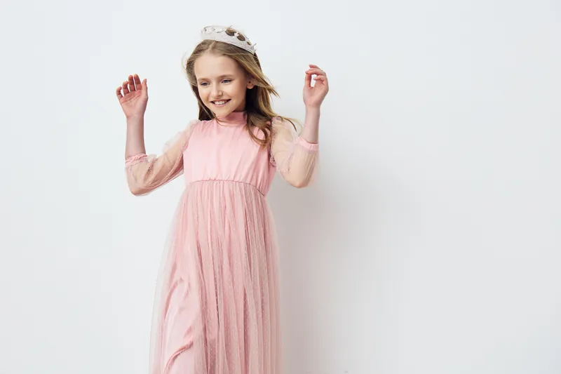 Joyful young girl in a pink dress celebrating with raised arms, wearing a crown against a minimalist white background, ideal for birthday or celebration themes