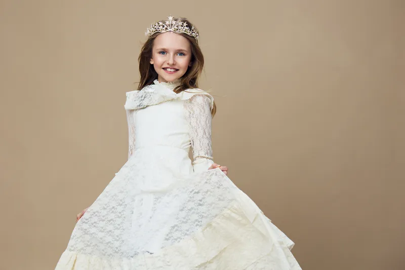 Young girl in an elegant white dress with a tiara, smiling joyfully against a soft beige background, capturing a sense of innocence and beauty