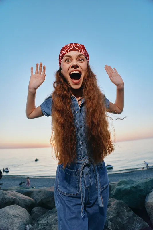 Excited woman wearing modern boho-western denim outfit and red bandana with long hair, captured in vibrant lifestyle film color against a beach sunset background.