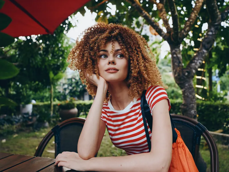 Portrait of a thoughtful young woman with curly hair sitting at an outdoor cafe, wearing a red and white striped shirt, relaxed posture, soft natural light, lush greenery.