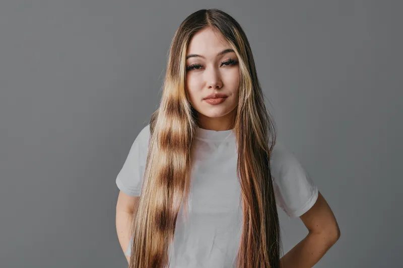 Woman with long straight hair wearing white t shirt standing against gray background looking at camera with relaxed expression and hands on hips in casual style.