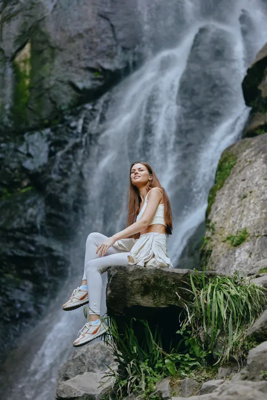 Young woman sitting on rock near waterfall in nature, wearing light sporty clothes and sneakers, enjoying peaceful outdoor scenery in lush green environment.