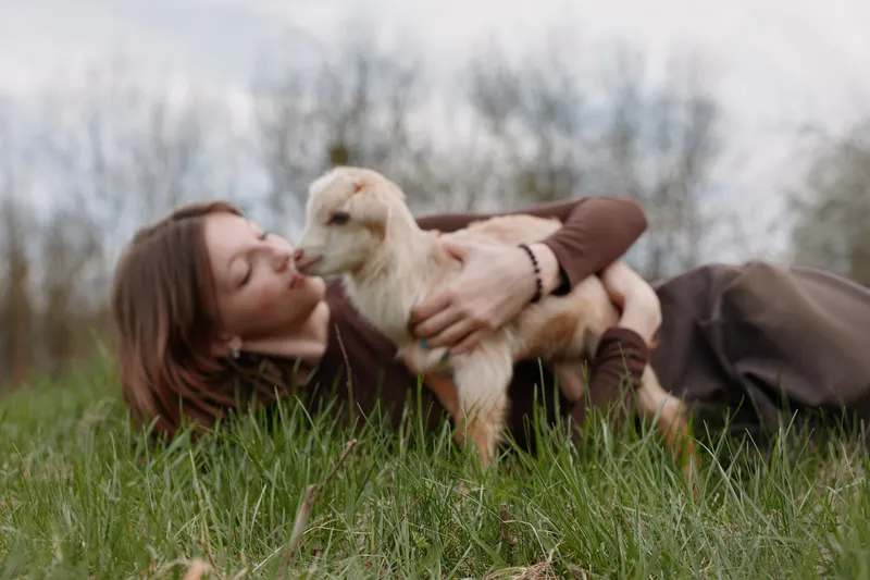 woman outdoors grass kiss affection nature countryside field lifestyle moment with goat kid in calm rural setting, tender bond between human and animal, warm light and peaceful atmosphere