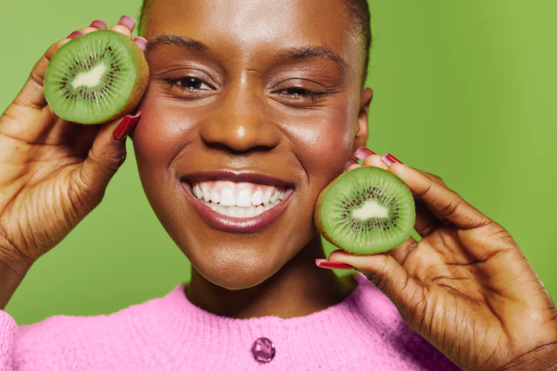 Happy African woman with dark skin holds two halves of kiwi fruit near face smiling brightly on green background. Concept of healthy lifestyle, fresh fruit, and natural beauty.