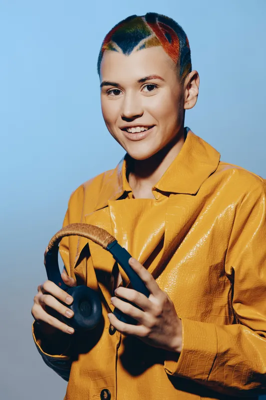 young woman with colorful short hair holding headphones, wearing a yellow jacket, smiling confidently against a soft blue background