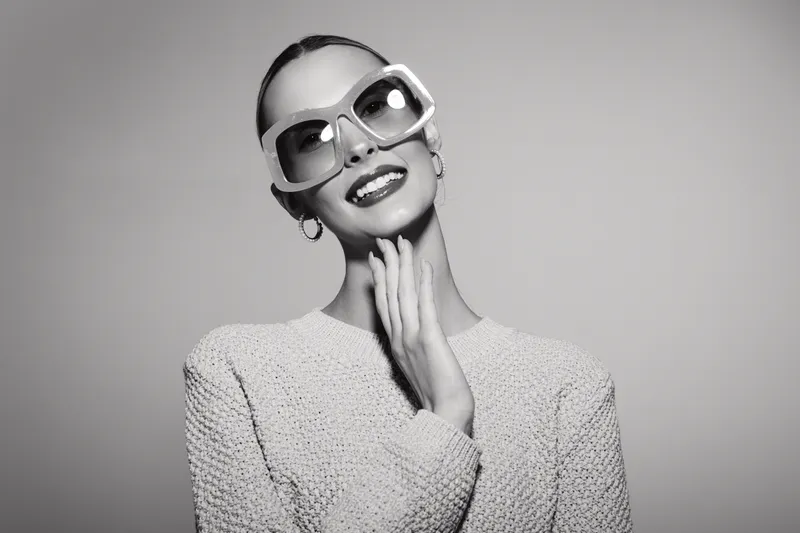 Elegant slim Asian woman with dark skin wearing large sunglasses and knitted sweater poses smiling in studio light background.