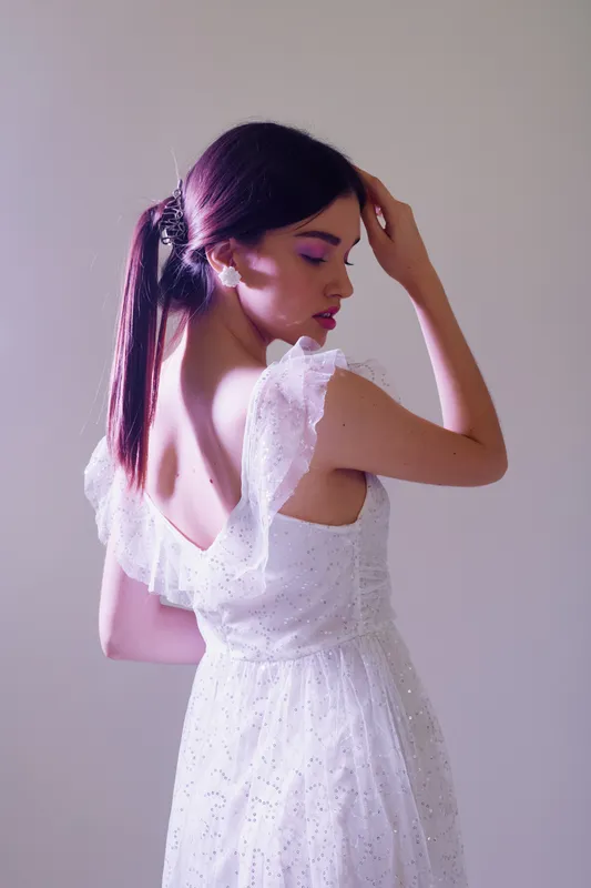 Young woman in a white lace dress poses with eyes closed and hand on head. Soft lighting highlights her beauty and fashion style. Studio shot on a neutral background.