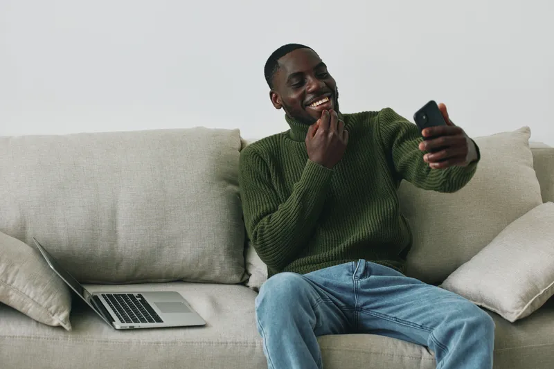 A smiling African American man enjoying a snack while video chatting on his smartphone, wearing a cozy green sweater, casual jeans, and sitting on a light colored sofa