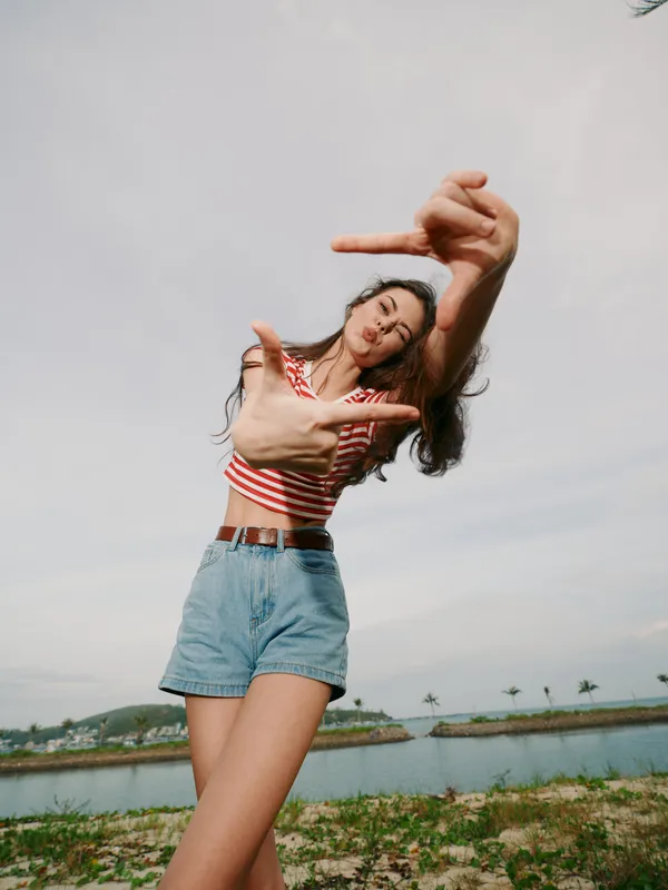 Young woman posing playfully outdoors in casual summer outfit, striped crop top and denim shorts, framing a dynamic hand gesture against a bright, open sky.