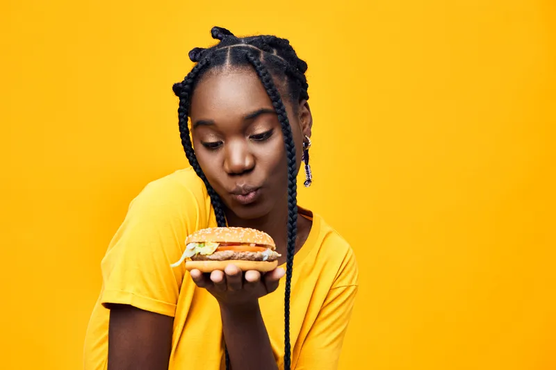 Young African woman with braids holding a colorful veggie burger, showcasing healthy eating and vibrant lifestyle against a bright yellow background