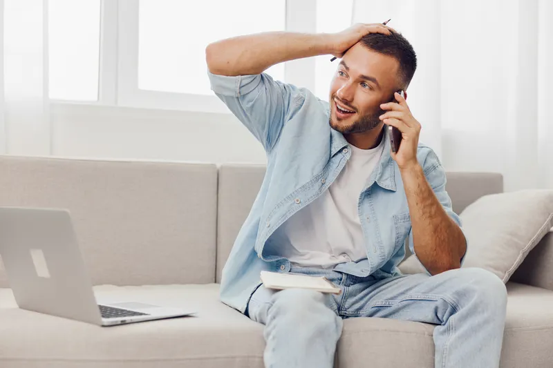 Excited man on a phone call, wearing casual attire. Bright living room setting with natural light creates a joyful atmosphere. Perfect for lifestyle themes.