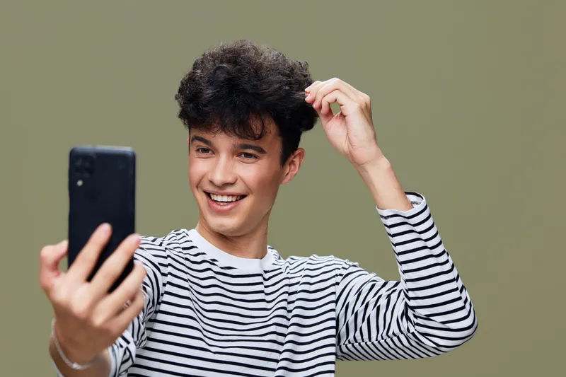 young man taking selfie with smartphone smiling, wearing striped shirt, curly hair, casual style, olive background, joyful expression