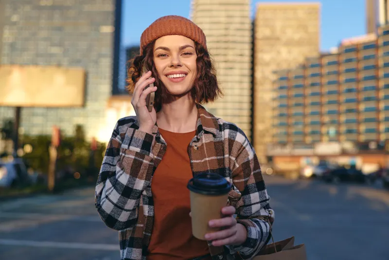 Woman smile on phone holding coffee in city sunlight, plaid jacket and beanie capture golden hour glow in candid lifestyle portrait emphasizing authenticity, emotional storytelling and mindful living