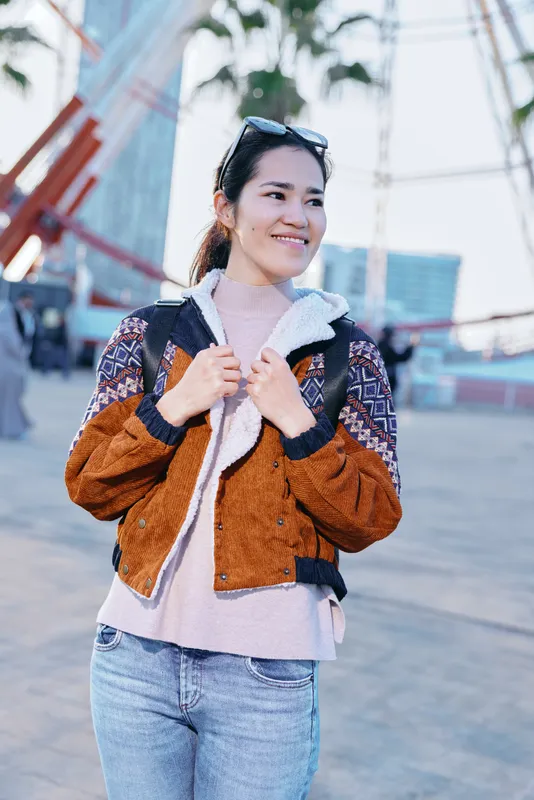 Smiling woman wearing a brown orange patterned jacket over a soft pink sweater, standing outdoors in an urban plaza, showcasing casual fashion, street style and an approachable vibe