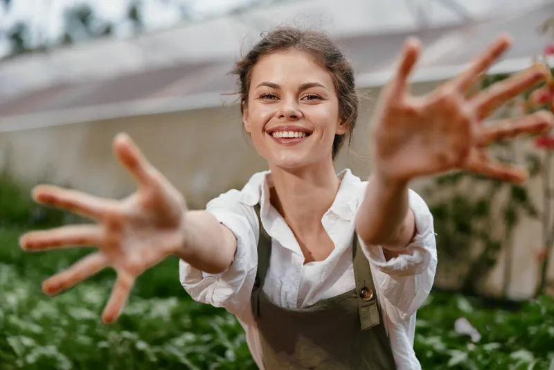 Smiling young woman in a garden, wearing an apron and reaching out, expressing joy in a vibrant green setting for healthy lifestyle concepts