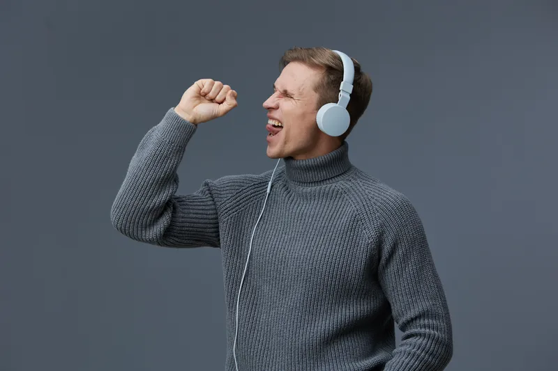 Happy young man wearing gray turtleneck sweater enjoying music with large white headphones on plain gray background, celebrating, feeling cheerful, relaxed, casual style, lifestyle concept